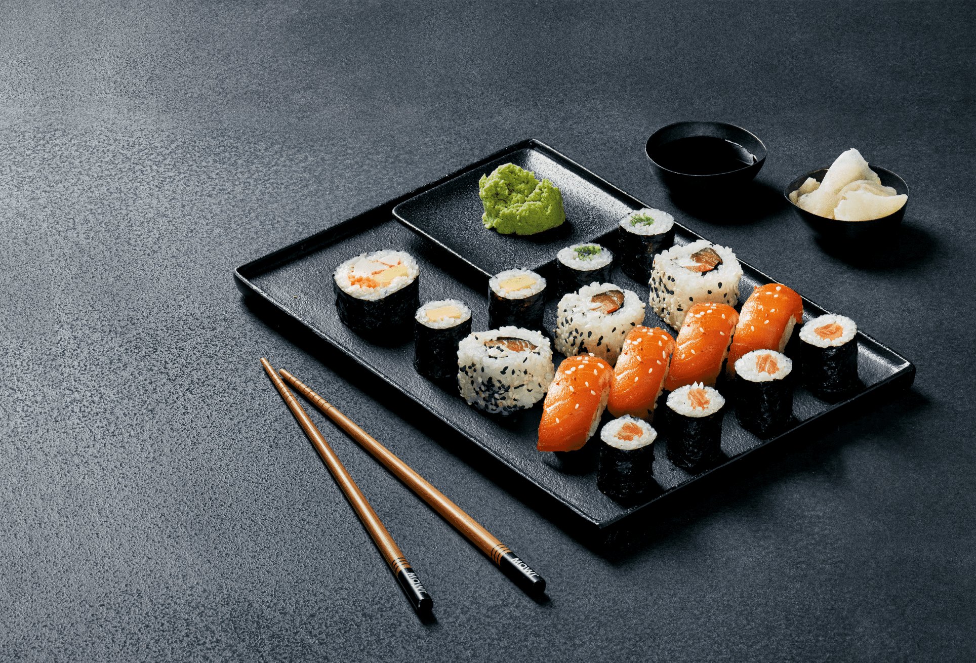 Zestaw różnorodnego sushi na czarnym półmisku z wasabi, sosem sojowym, imbirem i pałeczkami na ciemnym tle.