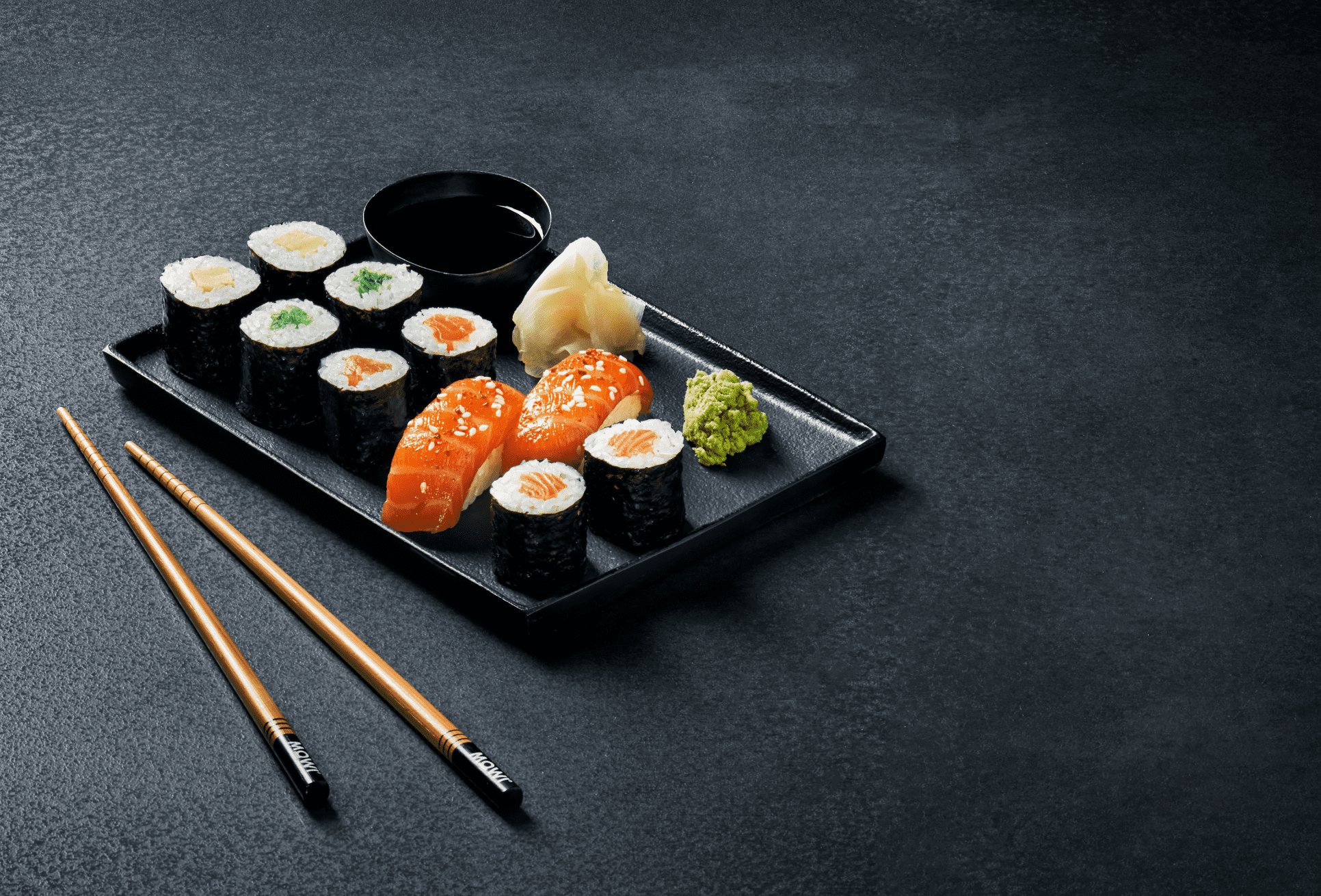Zestaw różnych rodzajów sushi, imbiru, wasabi i sosu sojowego na czarnej tacy z pałeczkami na ciemnym tle.