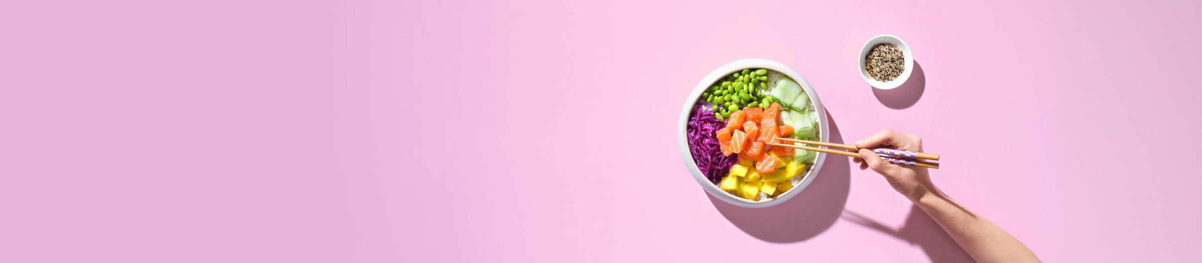Kolorowe poke bowl z łososiem, edamame, mango i kapustą na różowym tle, jedzone pałeczkami.