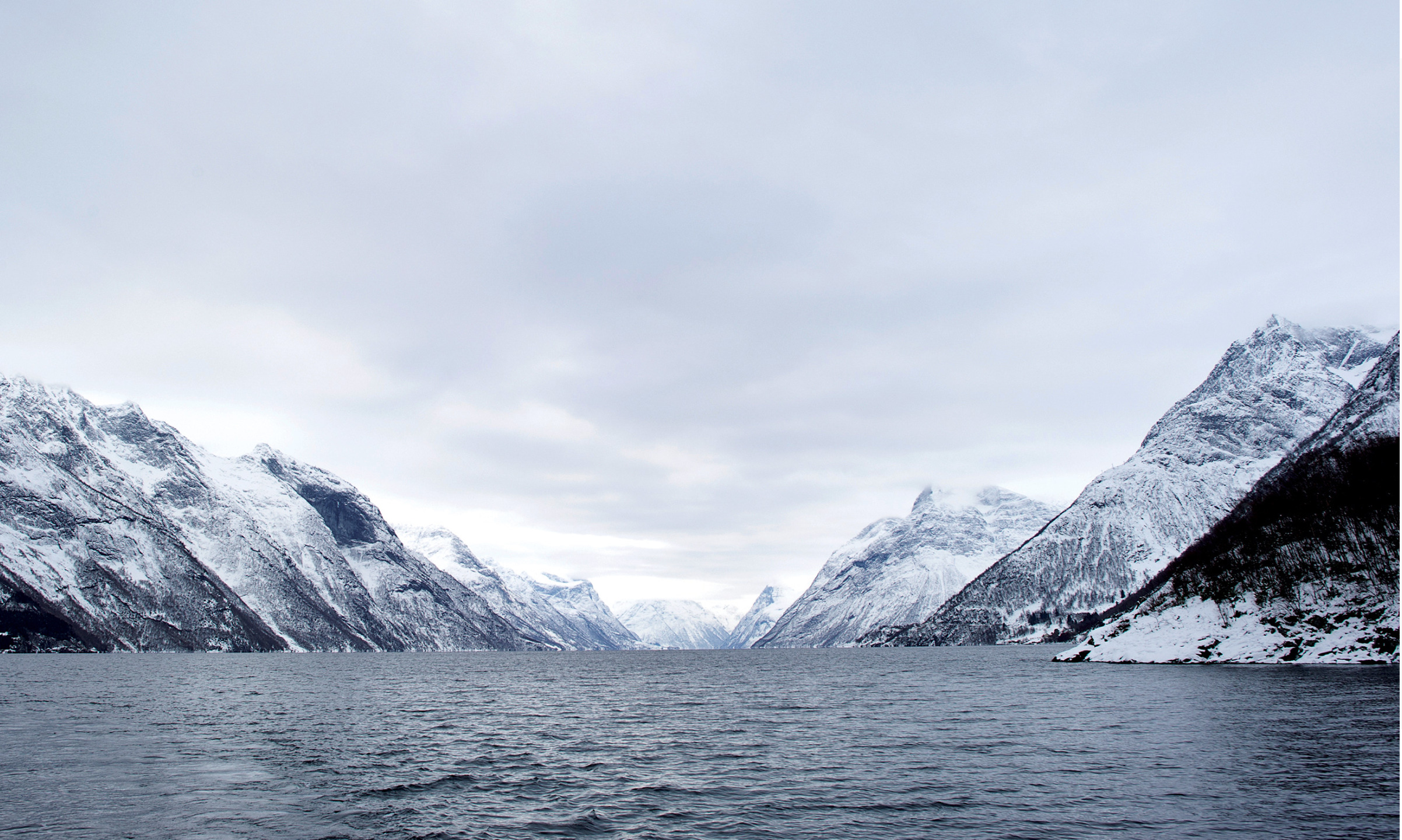 Norweski fiord otoczony wysokimi, ośnieżonymi górami pod zachmurzonym niebem.