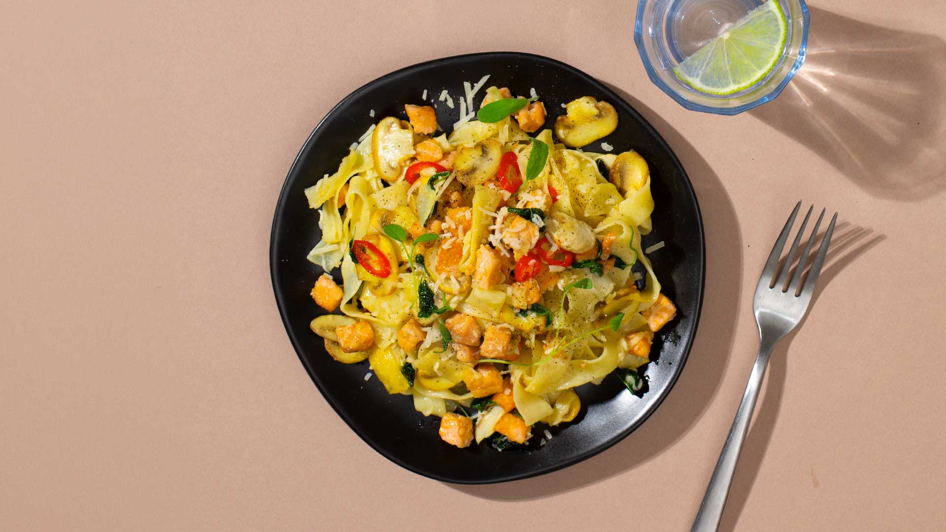 Talerz makaronu tagliatelle z łososiem, pieczarkami, chili i parmezanem, obok widelca i szklanki wody z limonką.