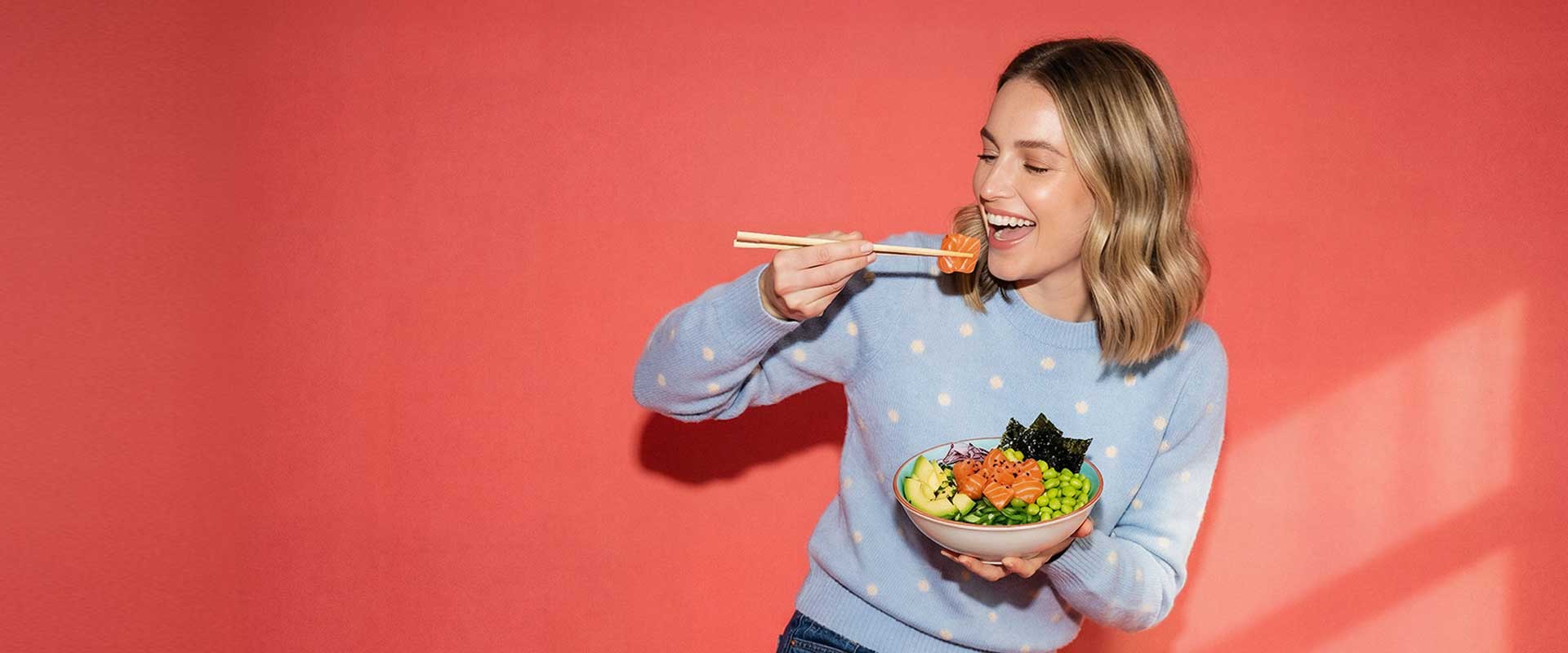 Uśmiechnięta blondynka w błękitnym swetrze je pałeczkami poke bowl z łososiem, awokado i edamame na koralowym tle.
