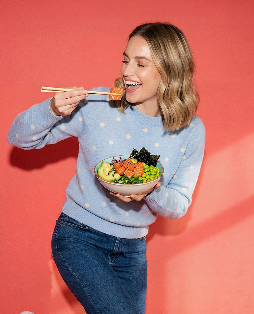Uśmiechnięta kobieta w jasnoniebieskim swetrze je pałeczkami kawałek łososia z miski pełnej składników poke bowl.